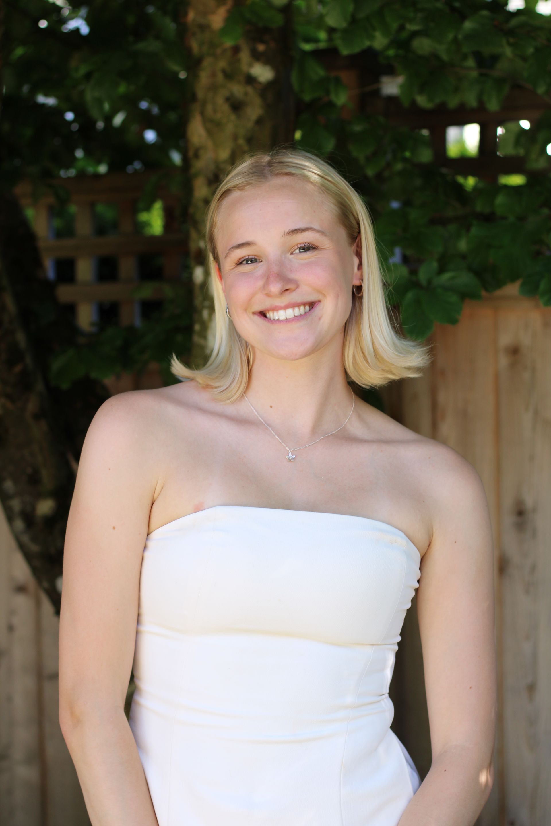 Person with blonde hair wearing a white strapless dress, smiling outdoors.