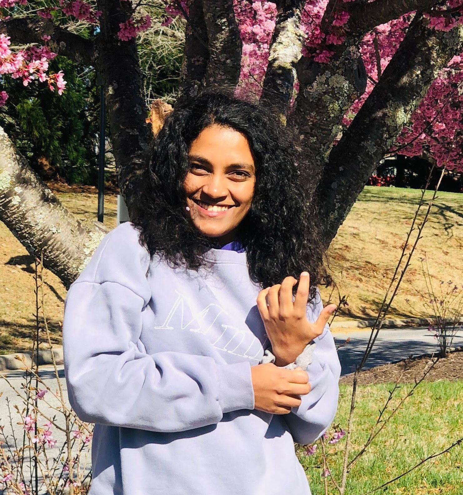 Person smiling, adjusting sleeve of a lavender sweatshirt, standing in front of a tree with pink blossoms.