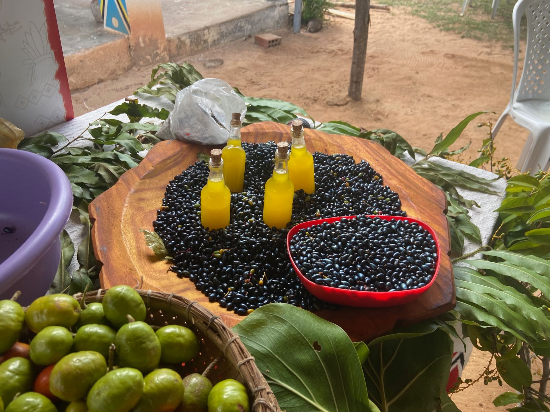 Bottles of yellow liquid atop black seeds on a wooden board; surrounded by green fruit and leaves.
