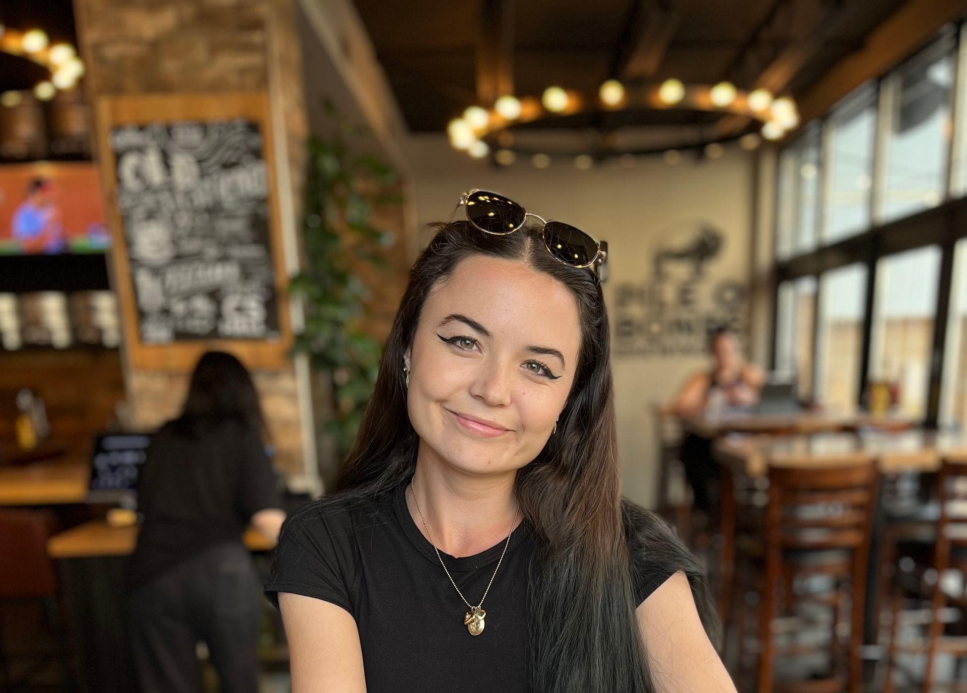 Person with sunglasses smiling indoors at a restaurant, surrounded by decor, other people visible.