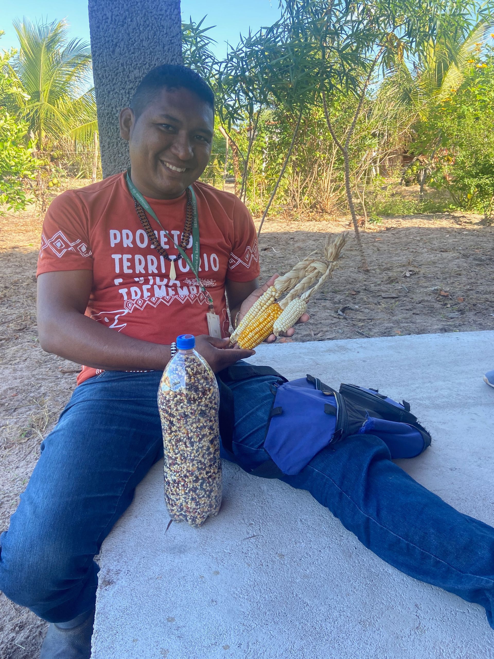 Person smiling, holding corn and a bottle of seeds, sitting outdoors.
