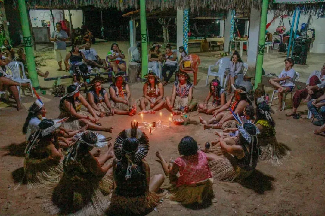People sit in a circle around candles. They wear traditional clothing. An outdoor gathering.