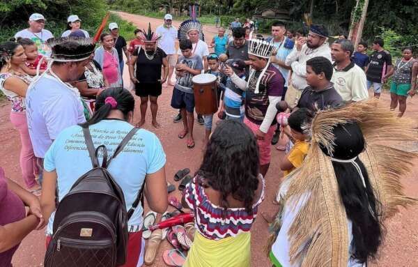 Group of people gathered outdoors, some holding a drum. Red dirt road, lush green trees.