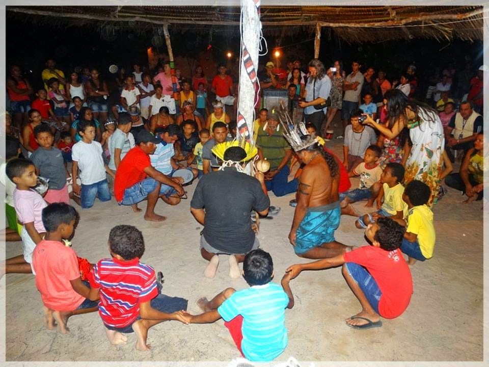 People gathered in a circle, some dancing, under a thatched roof at night.