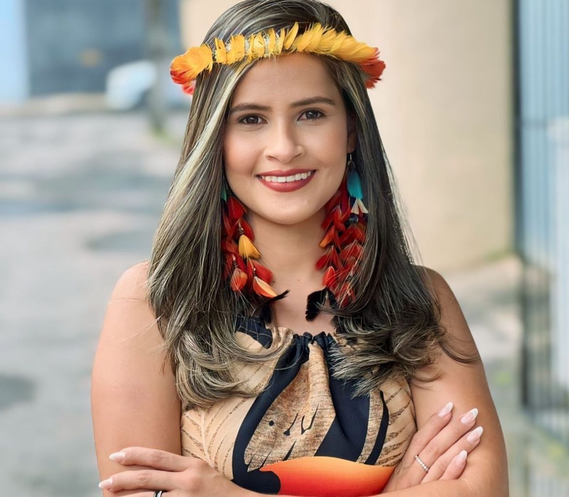 Person wearing a feathered headdress and earrings, smiling. She's outdoors in a patterned top.