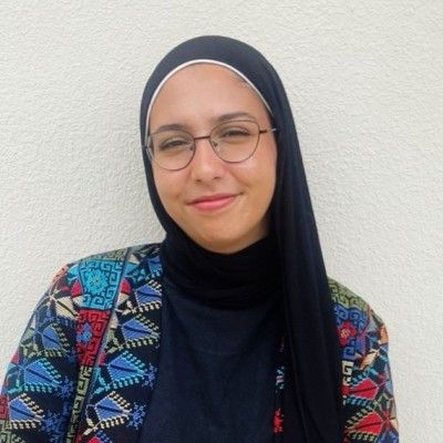 Person wearing glasses and a black headscarf, smiling, and colorful patterned jacket.