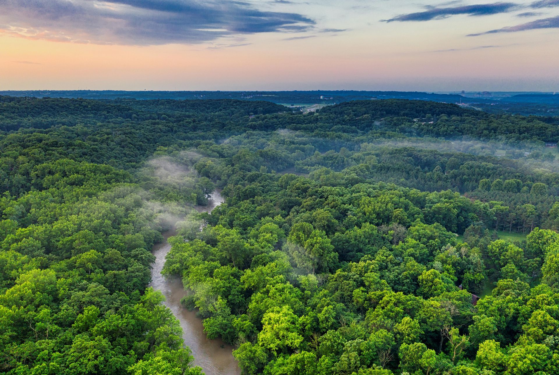 Aerial view of a lush green forest with a winding river and a misty atmosphere under a colorful sky.