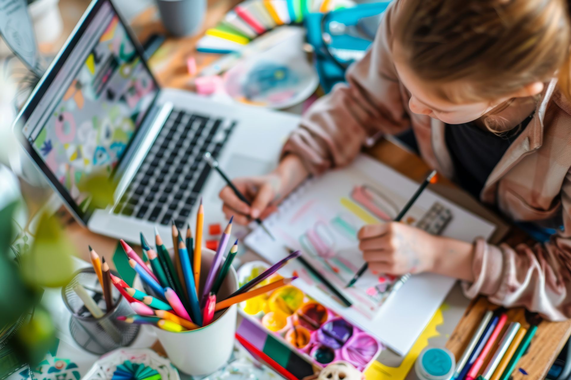 A woman is sitting at a table with a laptop and pencils.