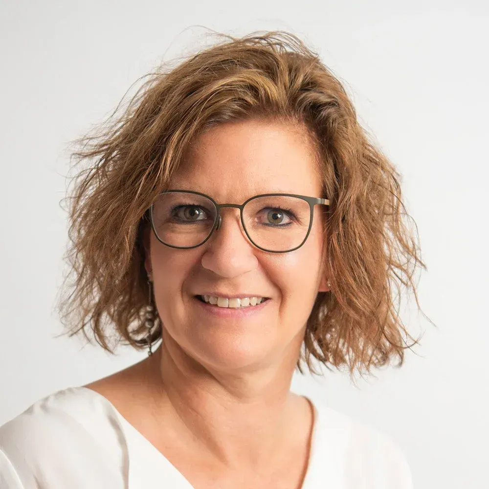 Frau mit Brille lächelt in die Kamera. Sie hat kurzes, hellbraunes Haar und trägt ein weißes Oberteil.