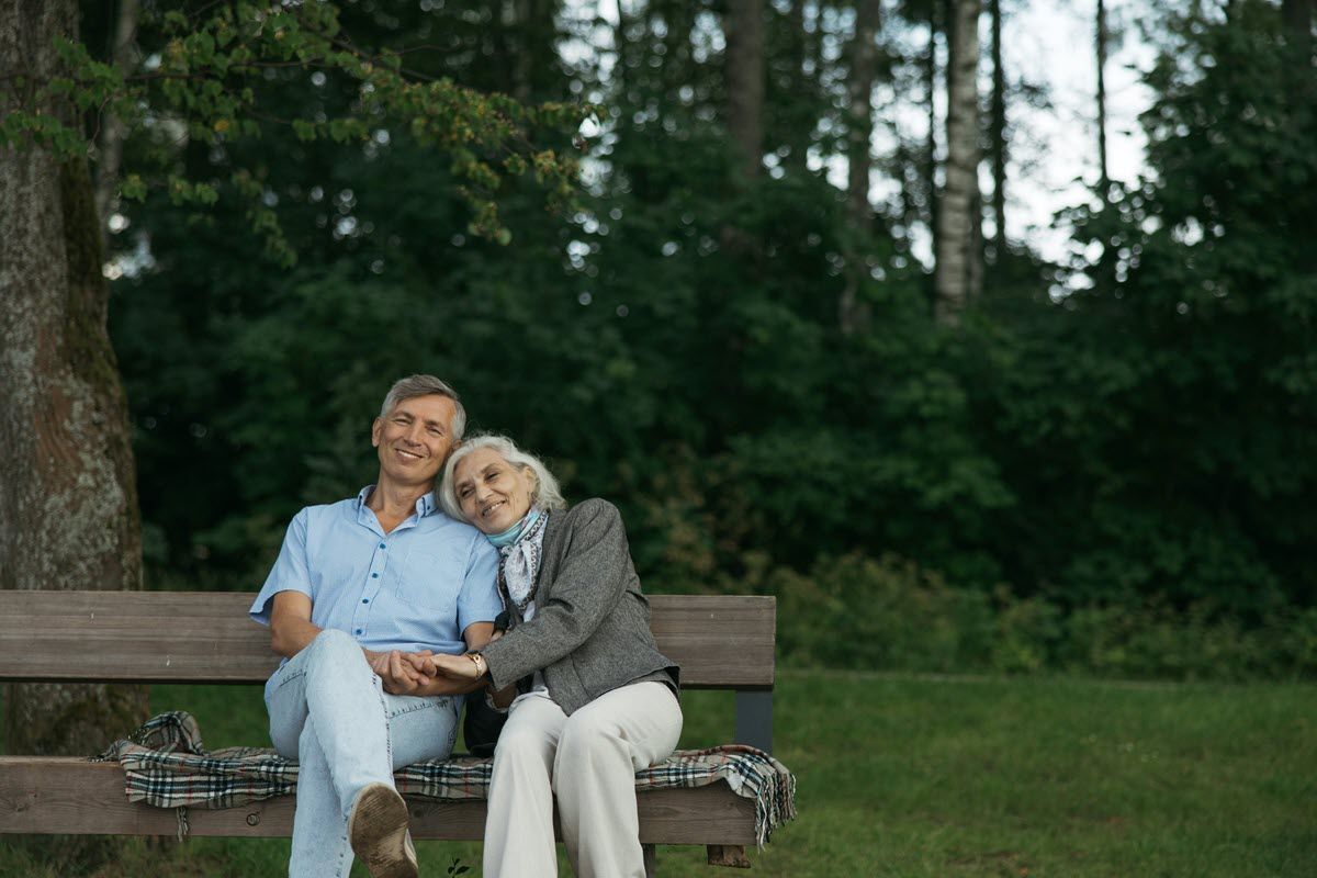 Couple sitting on park bench, holding hands. Man smiles, woman rests head on his shoulder. Forest background.