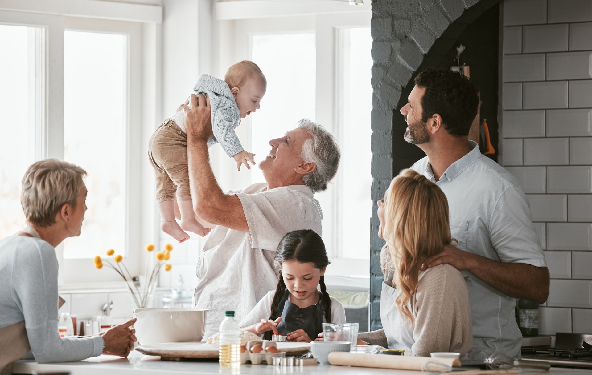 Family in a kitchen: grandfather holding baby, daughter baking, others watching and smiling.