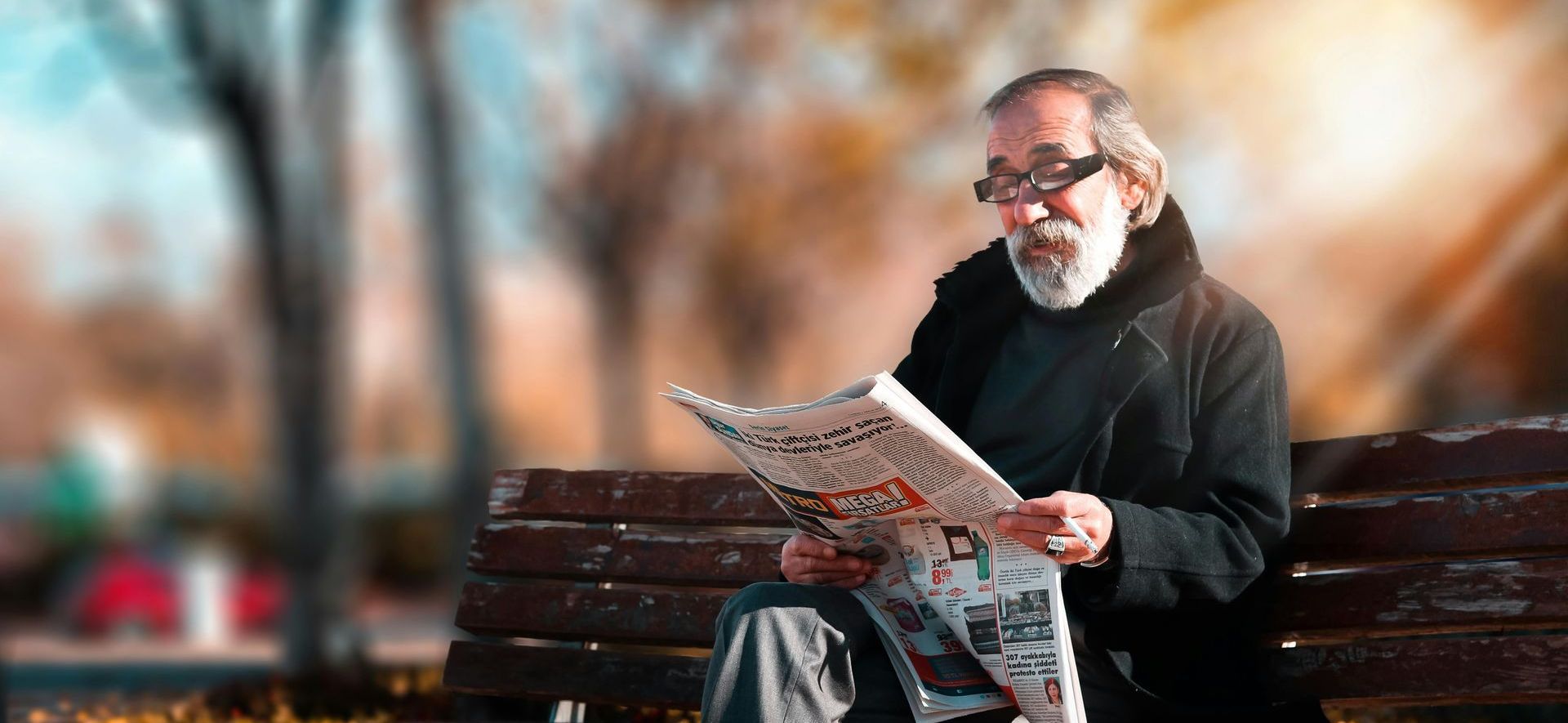Older man reading a newspaper, seated on a park bench in autumn. Sunlight shines on his face and the foliage.