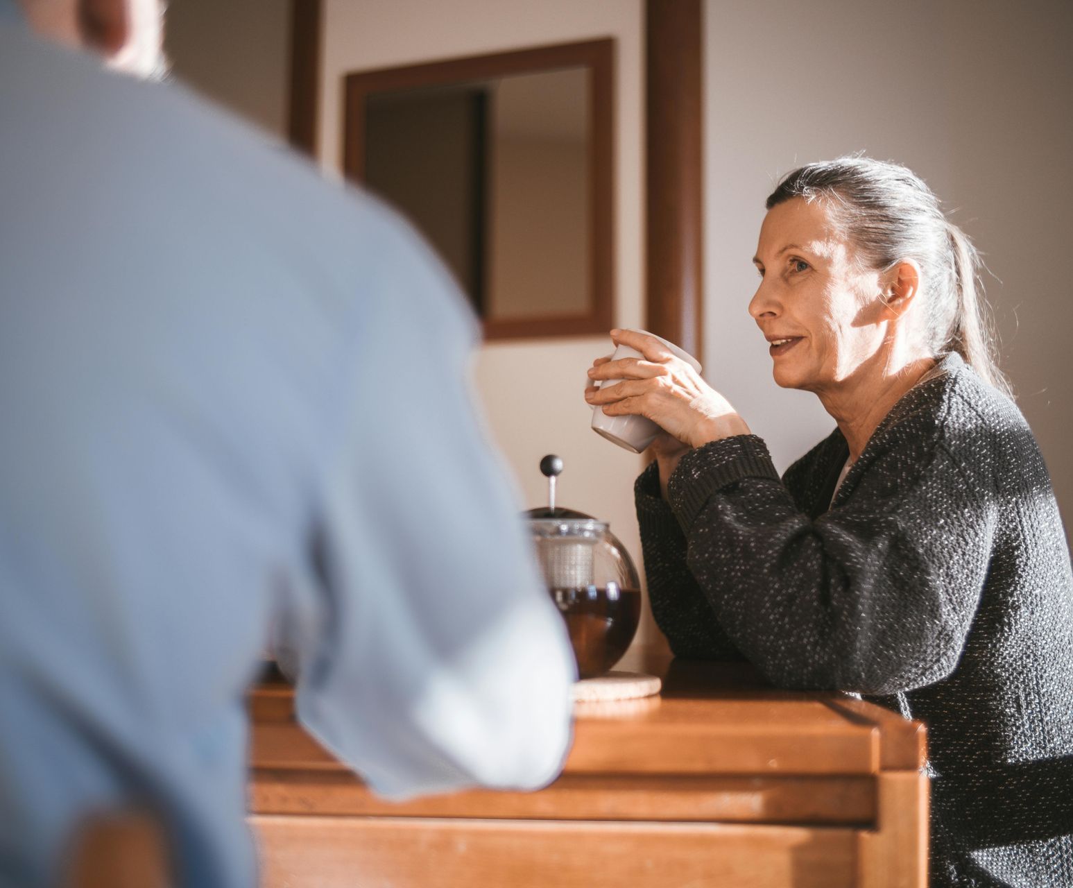 Woman, with a cup, talking to a man across a wooden table; teapot and mirror in background.