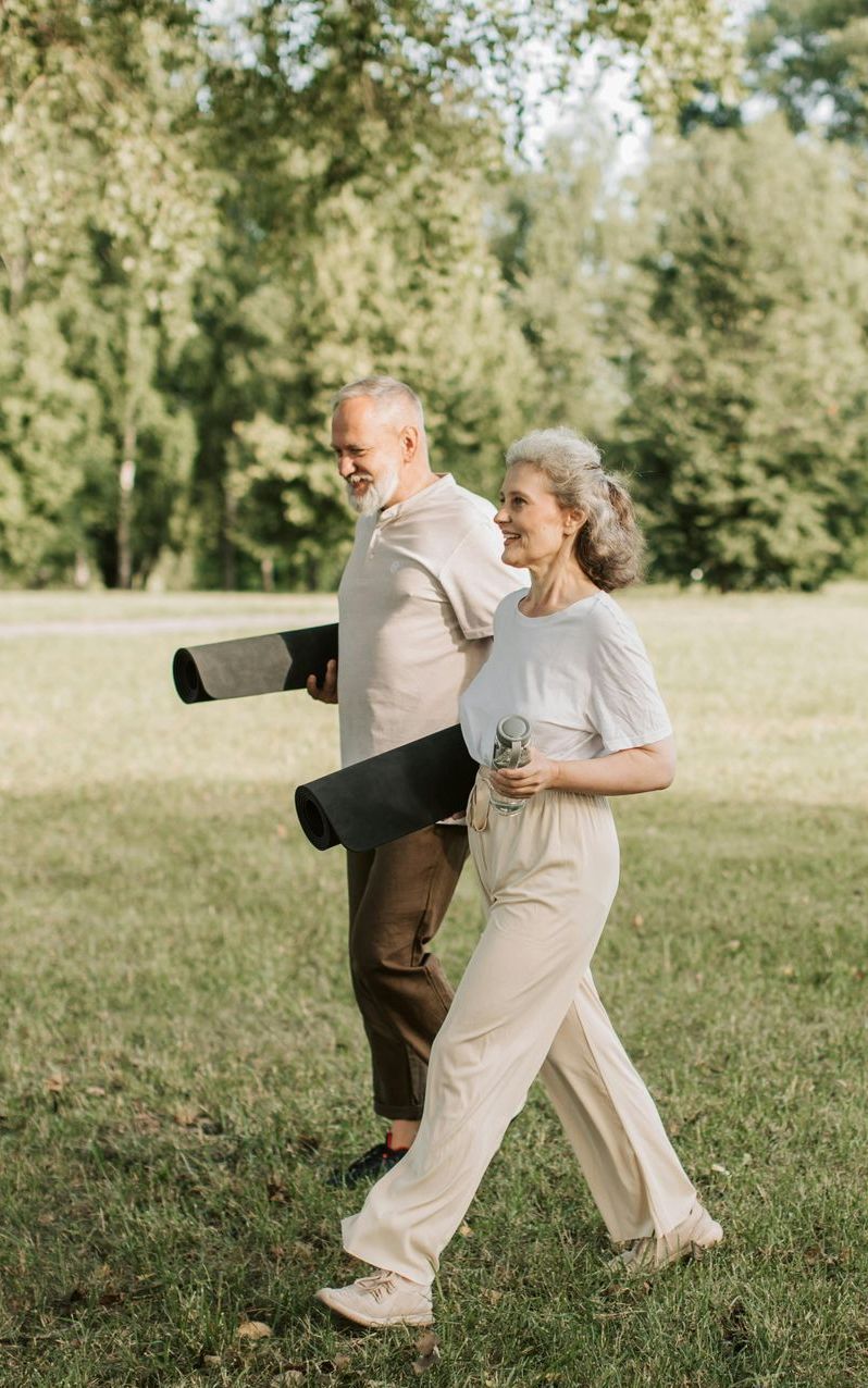 Older couple walking in a park, holding yoga mats. Green grass, trees, smiling faces.