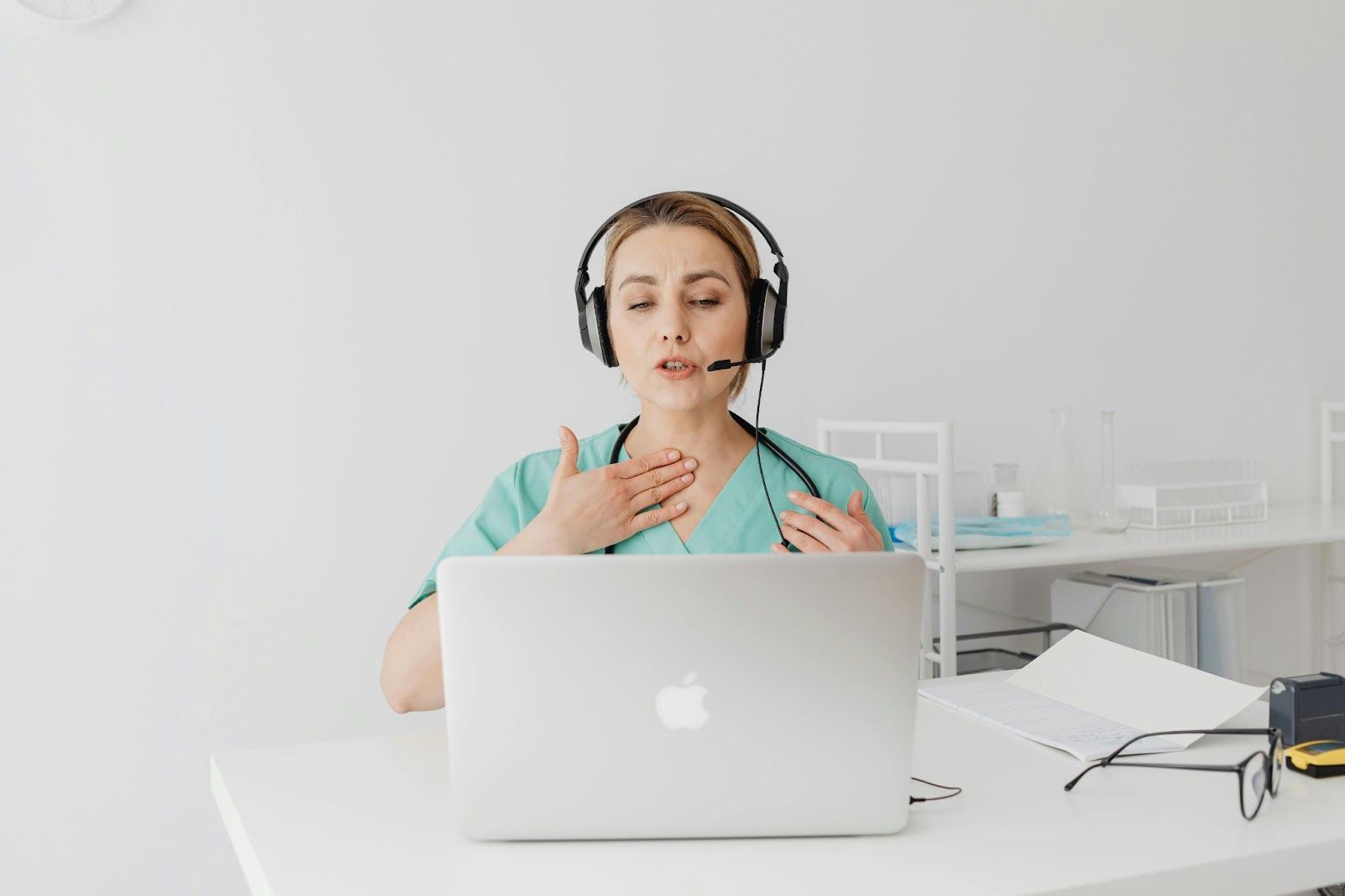 A healthcare professional in scrubs and a headset consults via laptop, touching their neck.