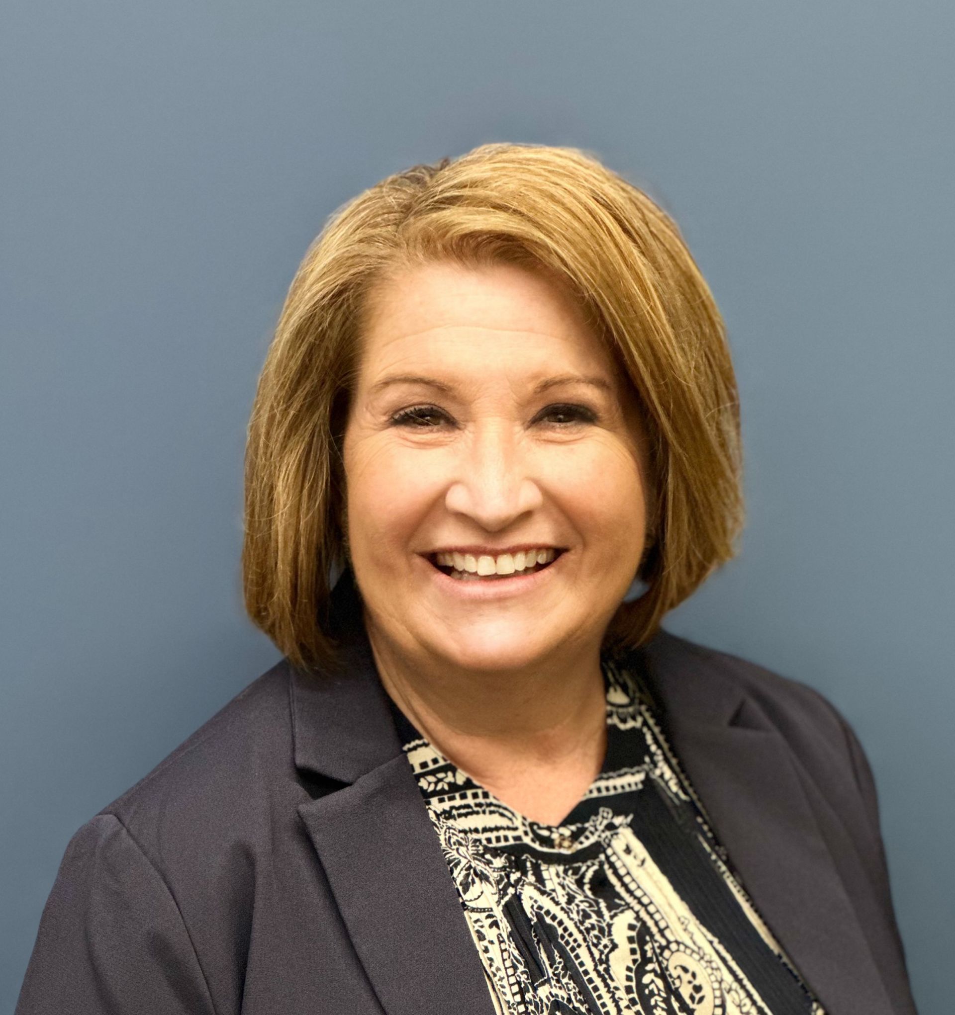 A woman wearing a blazer and a paisley shirt is smiling for the camera.