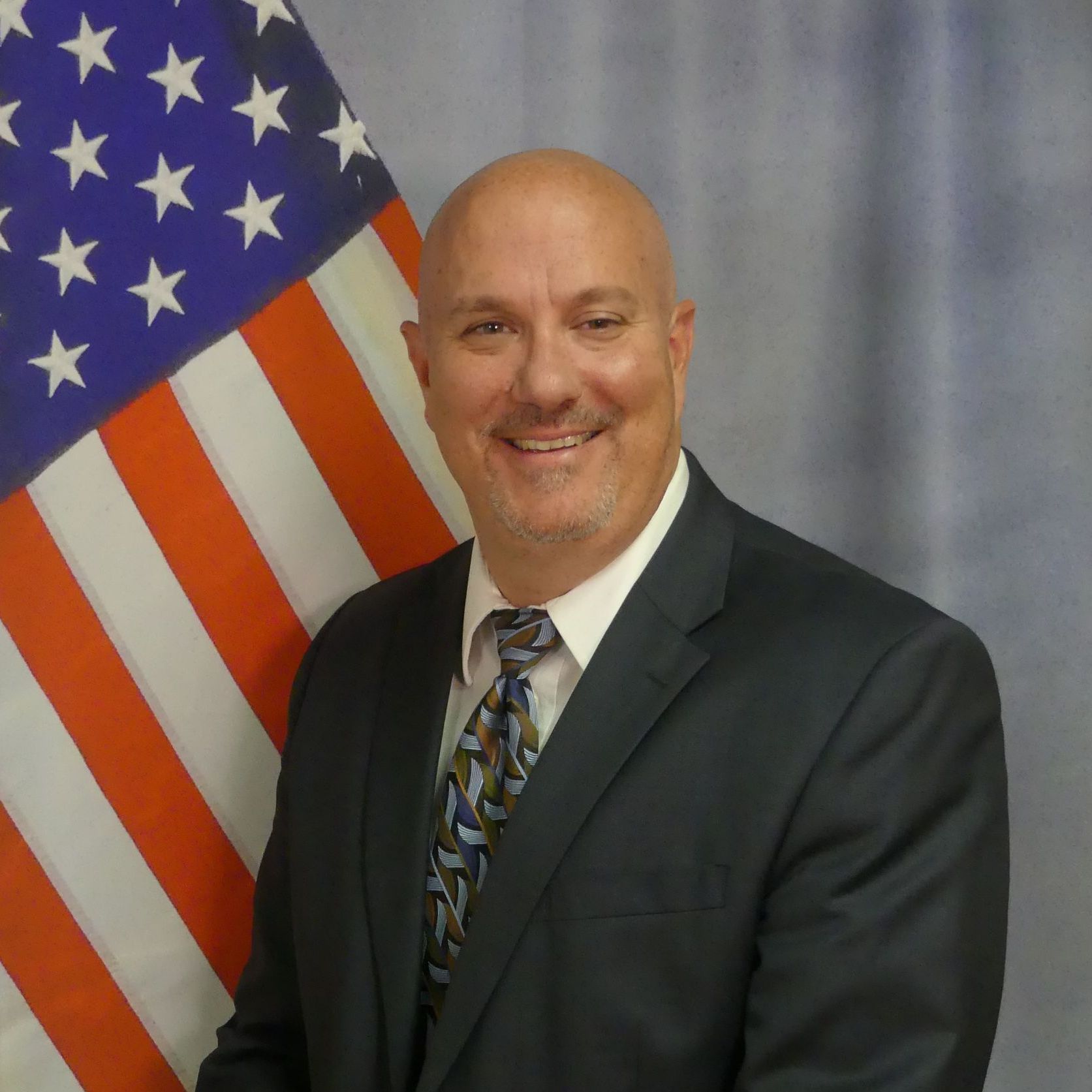 A man in a suit and tie stands in front of an american flag