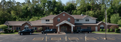 A one-story brick building with a covered entrance, several parked cars, and a surrounding parking lot and trees.
