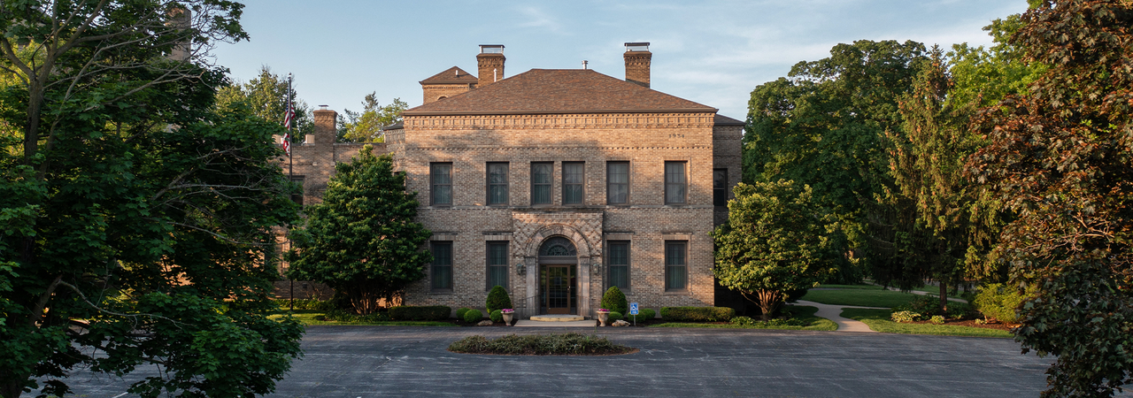 A grand stone mansion with a gray roof is surrounded by trees in the daytime.