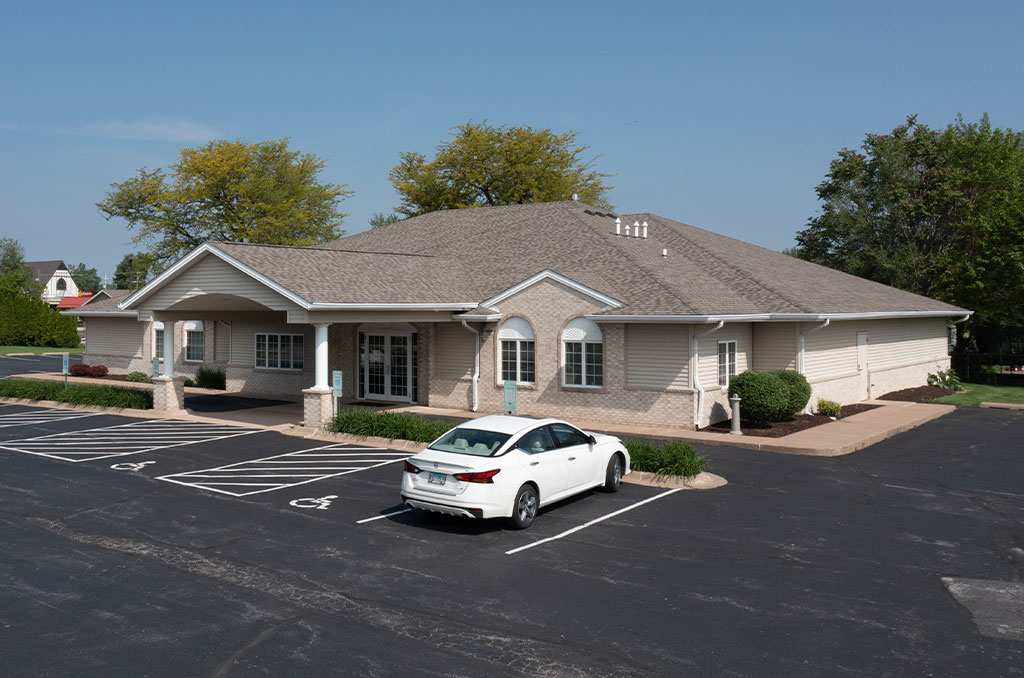 Exterior of a single-story building with a white car parked in front; blue sky and trees in the background.