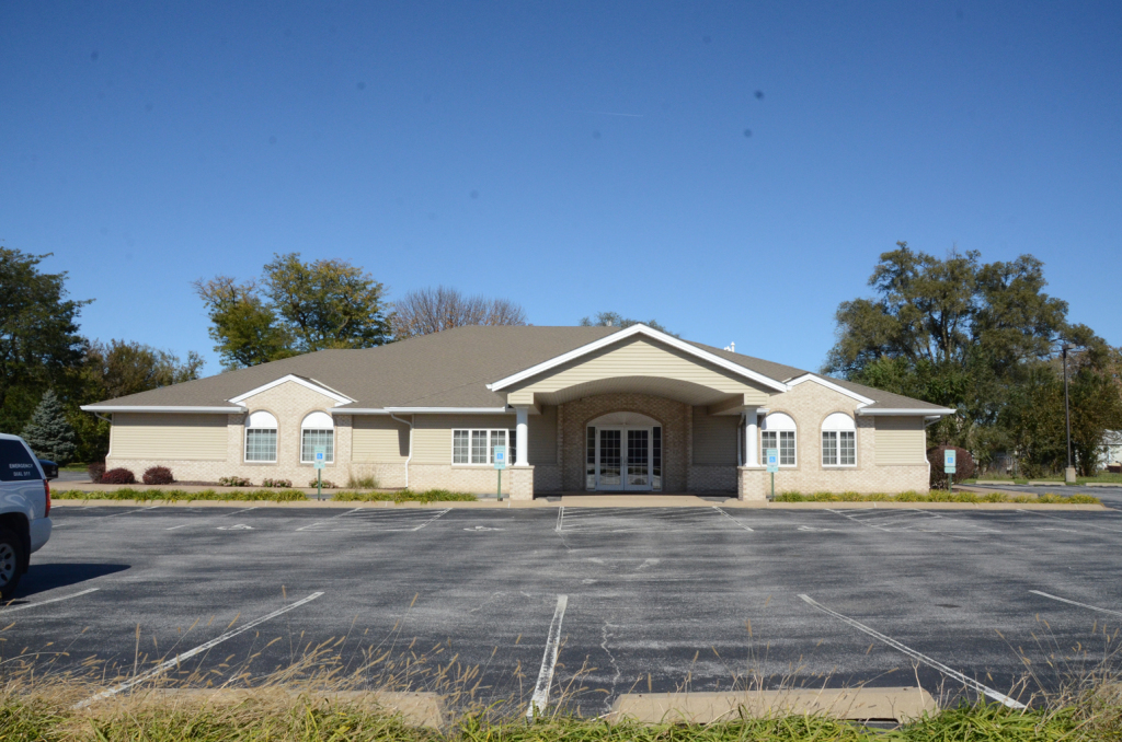 Tan, one-story building with a light-colored roof and a parking lot in front under a blue sky.