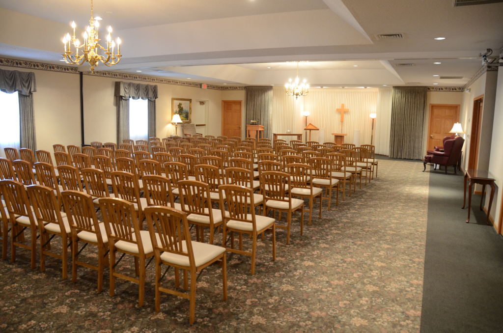 Funeral home chapel with rows of chairs facing an altar; ornate chandeliers and carpeted floor.