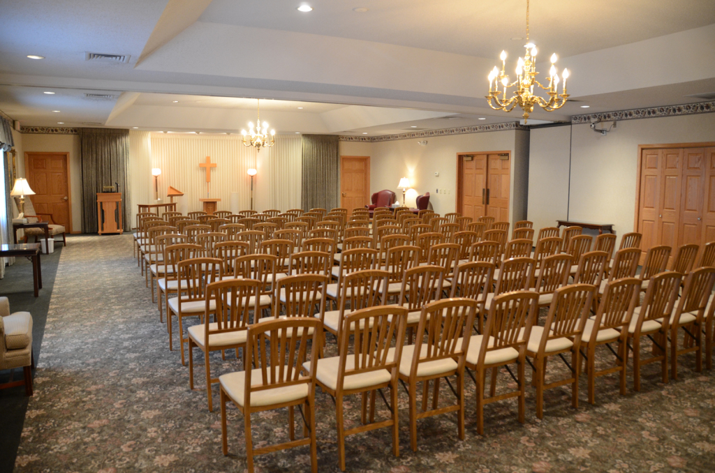 Rows of empty wooden chairs in a beige-toned room, likely a funeral home chapel, with a chandelier.