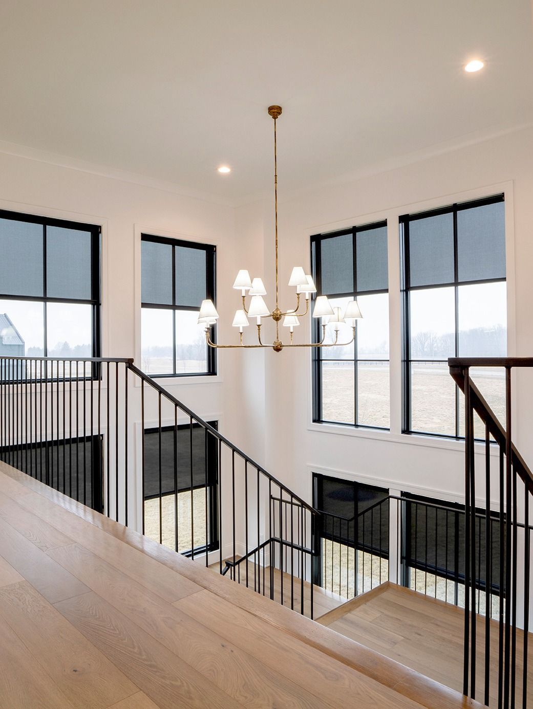 Interior staircase with black-framed windows with shades, a chandelier, and a wood banister.