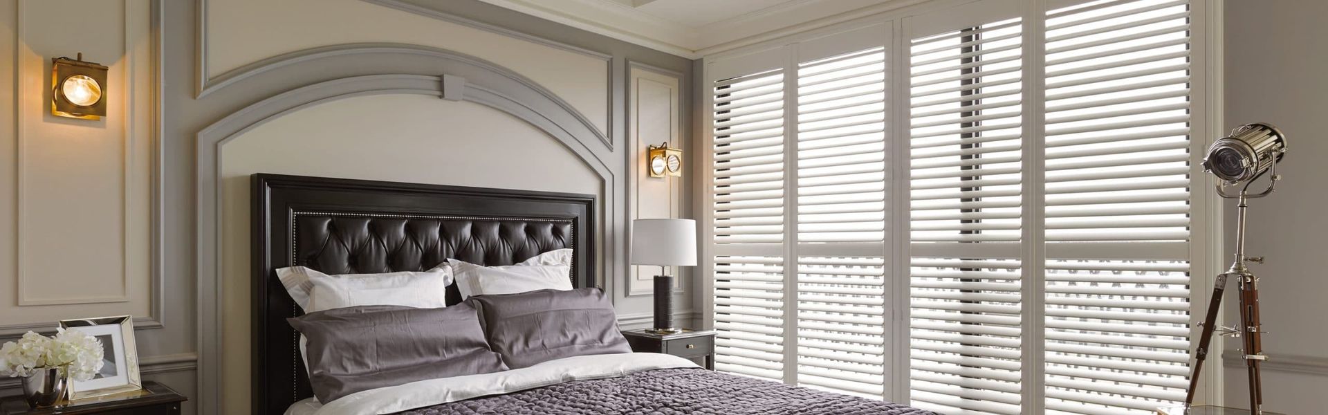 Bedroom with bed, windows with blinds, lamps, and decorative wall paneling.