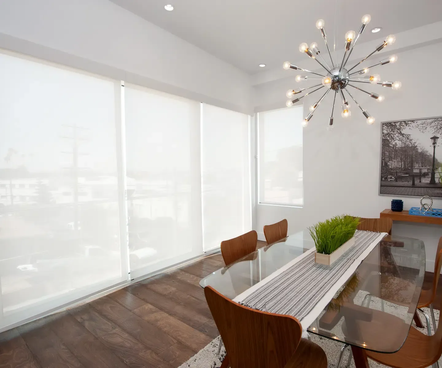 Dining room with glass table, wooden chairs, and roller shades.