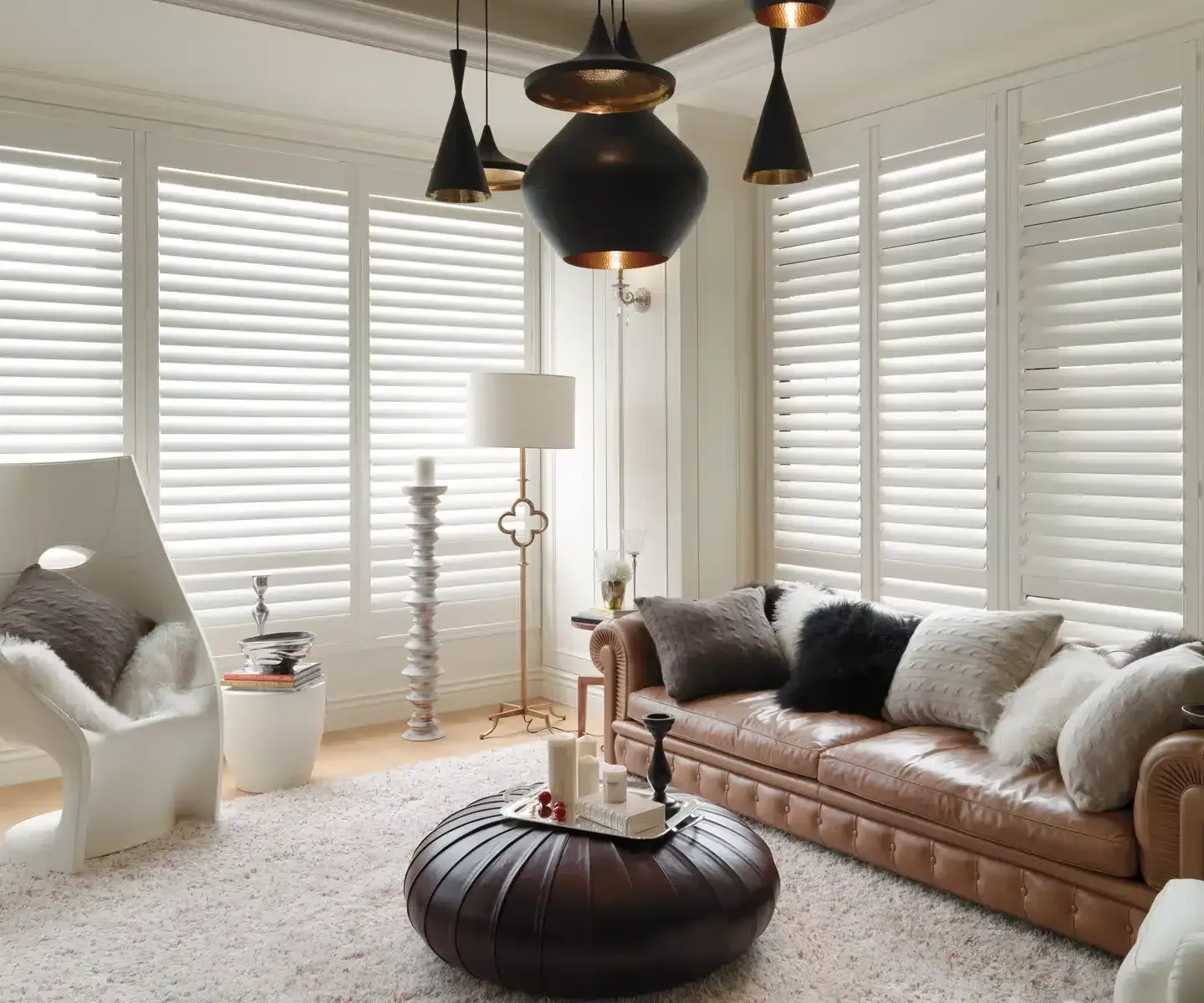 Living room with white shutters, leather couch, ottoman, and modern light fixtures.