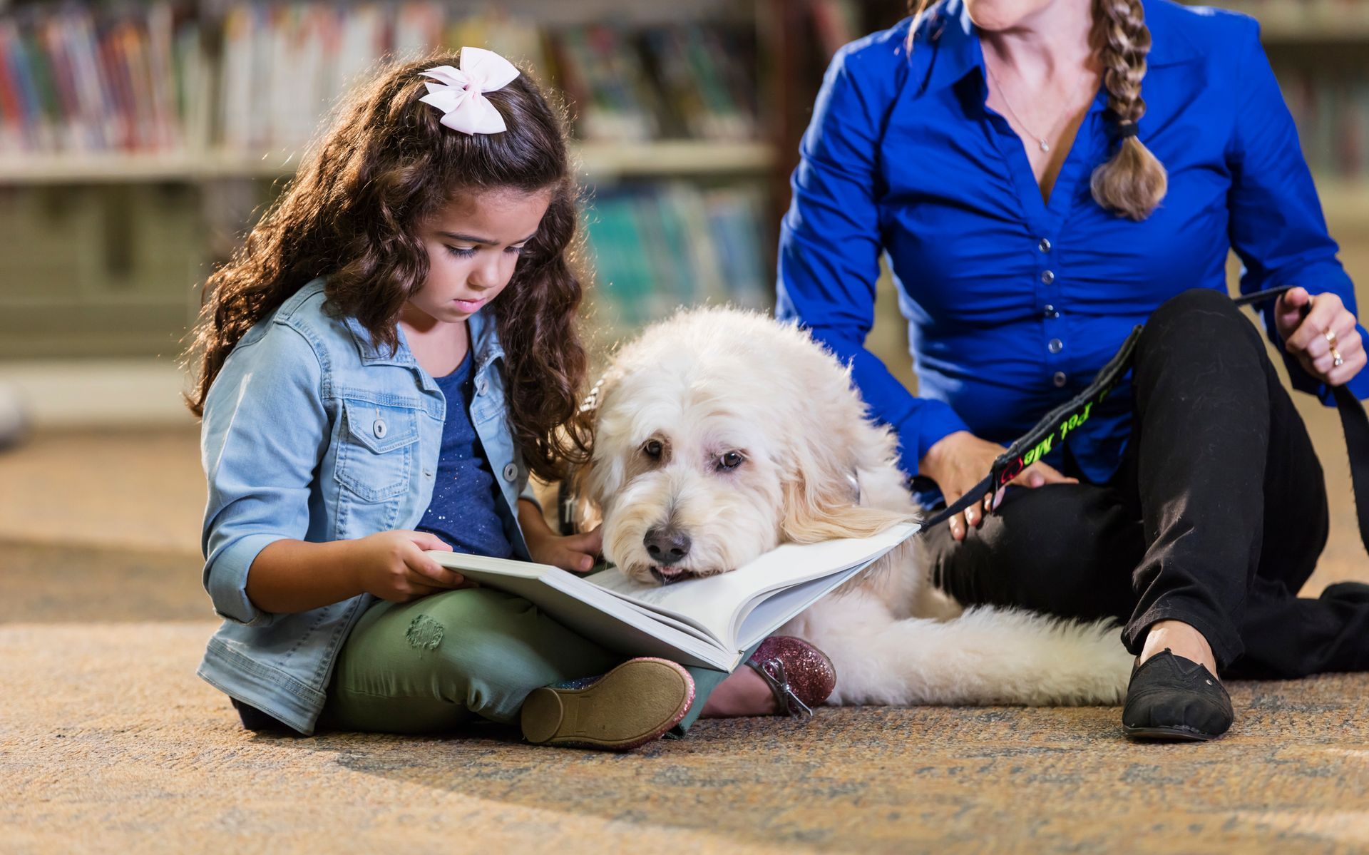 A woman and a little girl are sitting on the floor reading a book to a dog.
