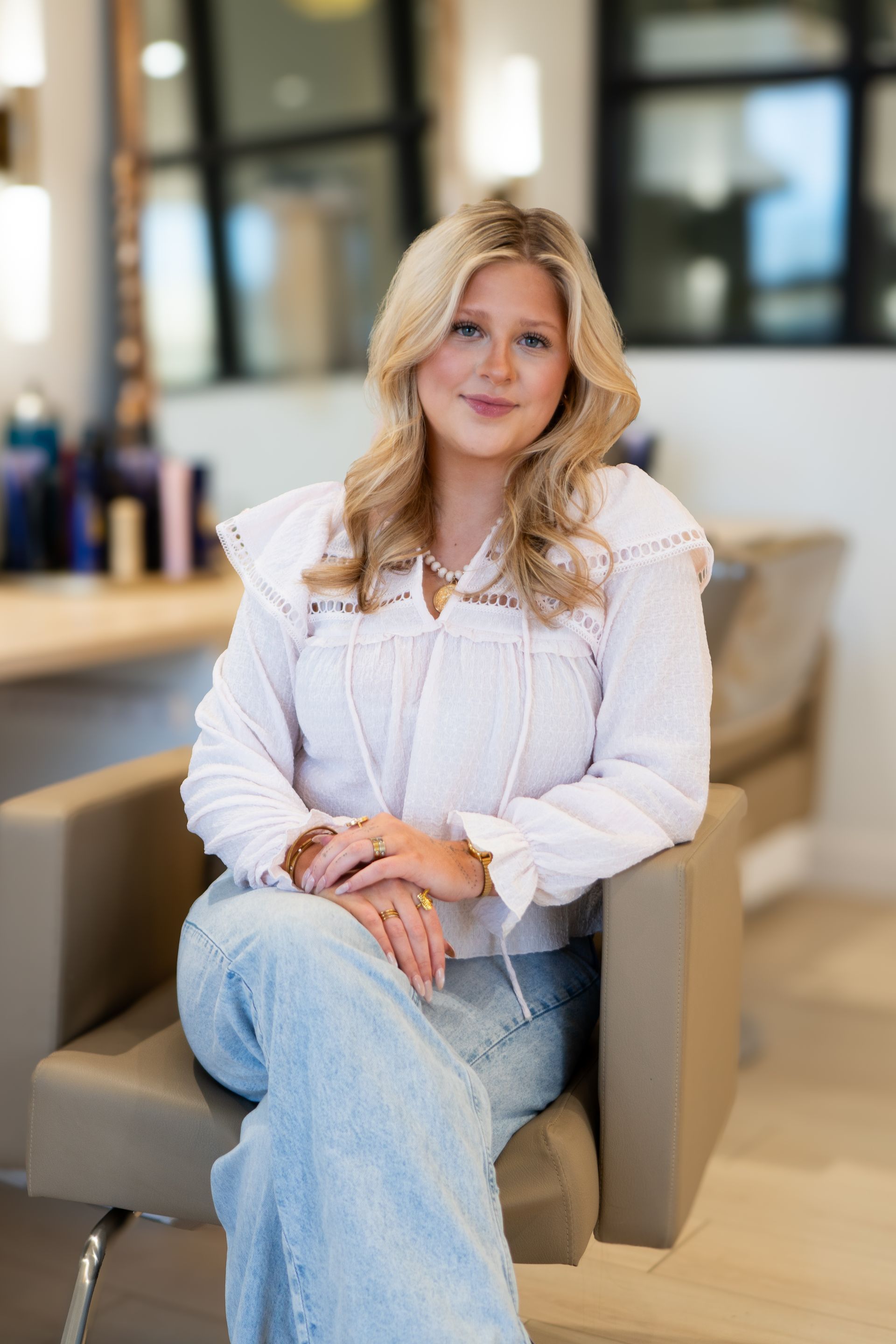 Woman with blonde hair sits in salon chair, wearing white top and jeans.