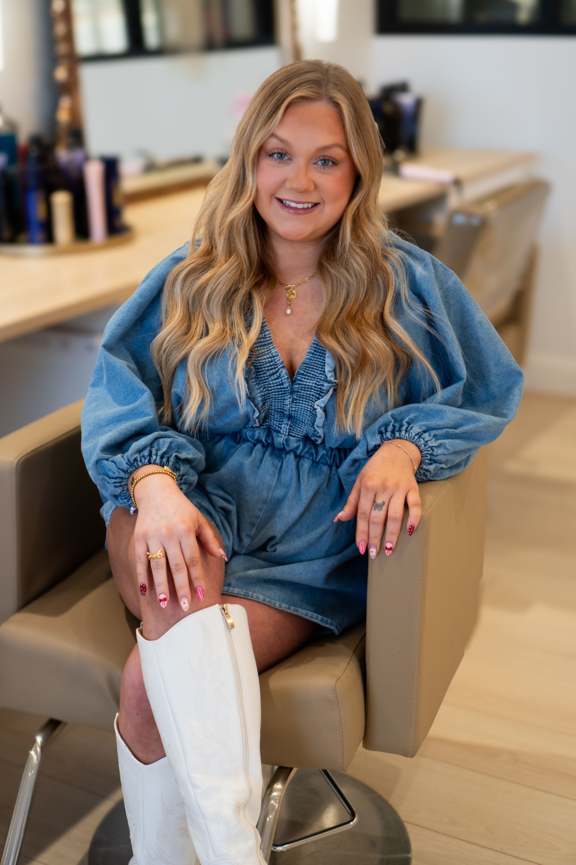 Woman in denim jumpsuit and white boots sits in a salon chair, smiling.