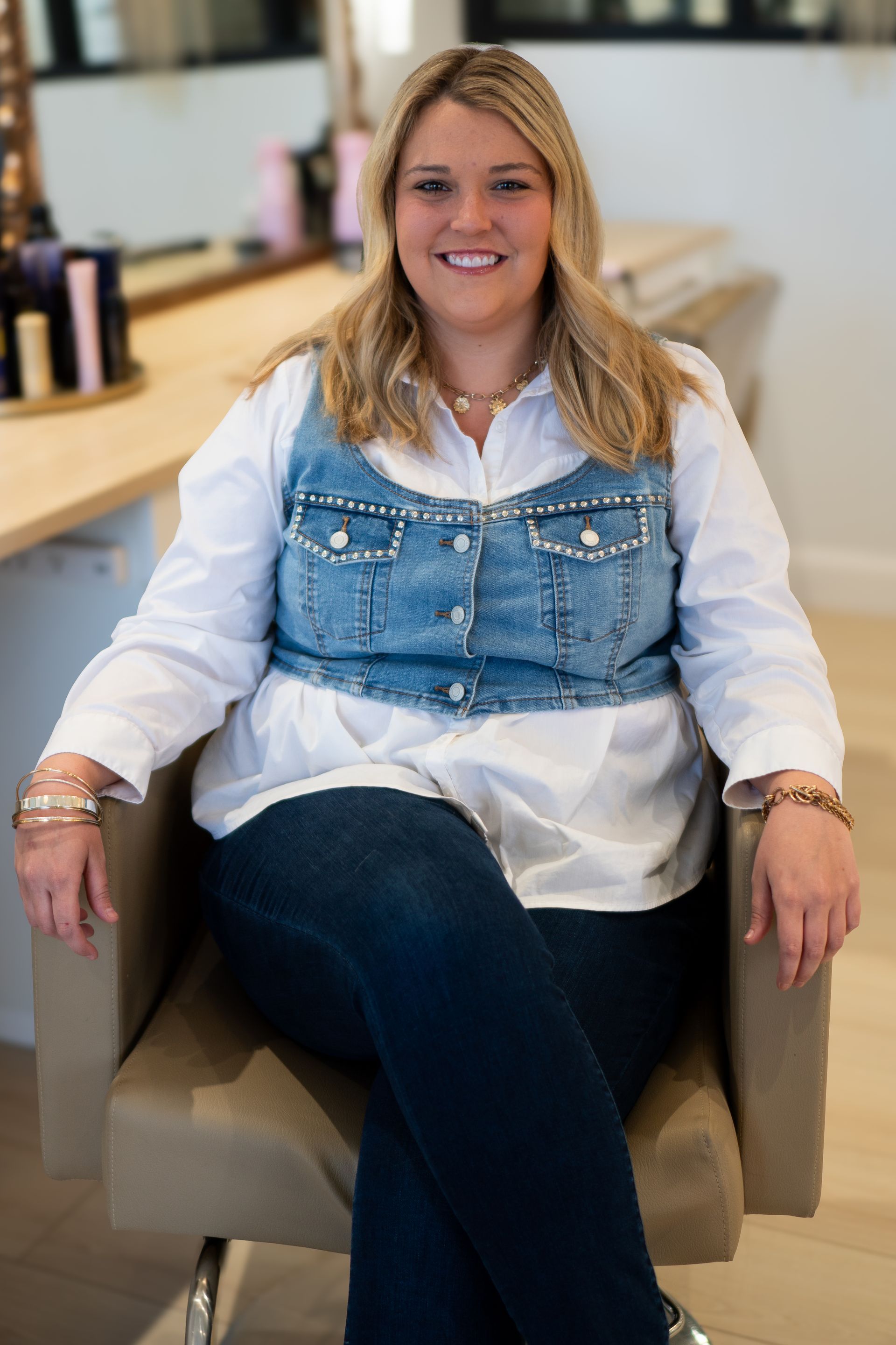Woman in denim vest and white shirt smiles while seated in a salon chair.