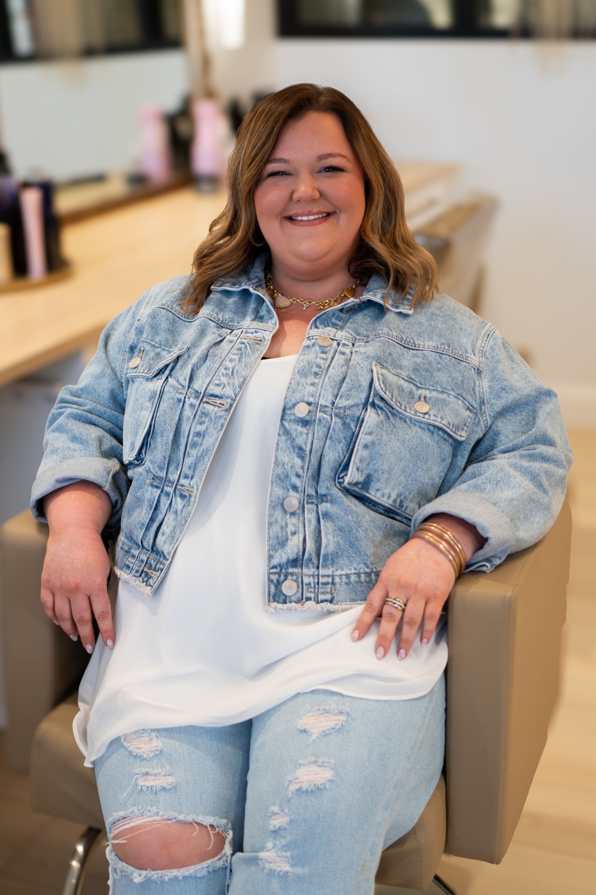 Woman seated, smiling, wearing denim jacket and jeans in a salon.