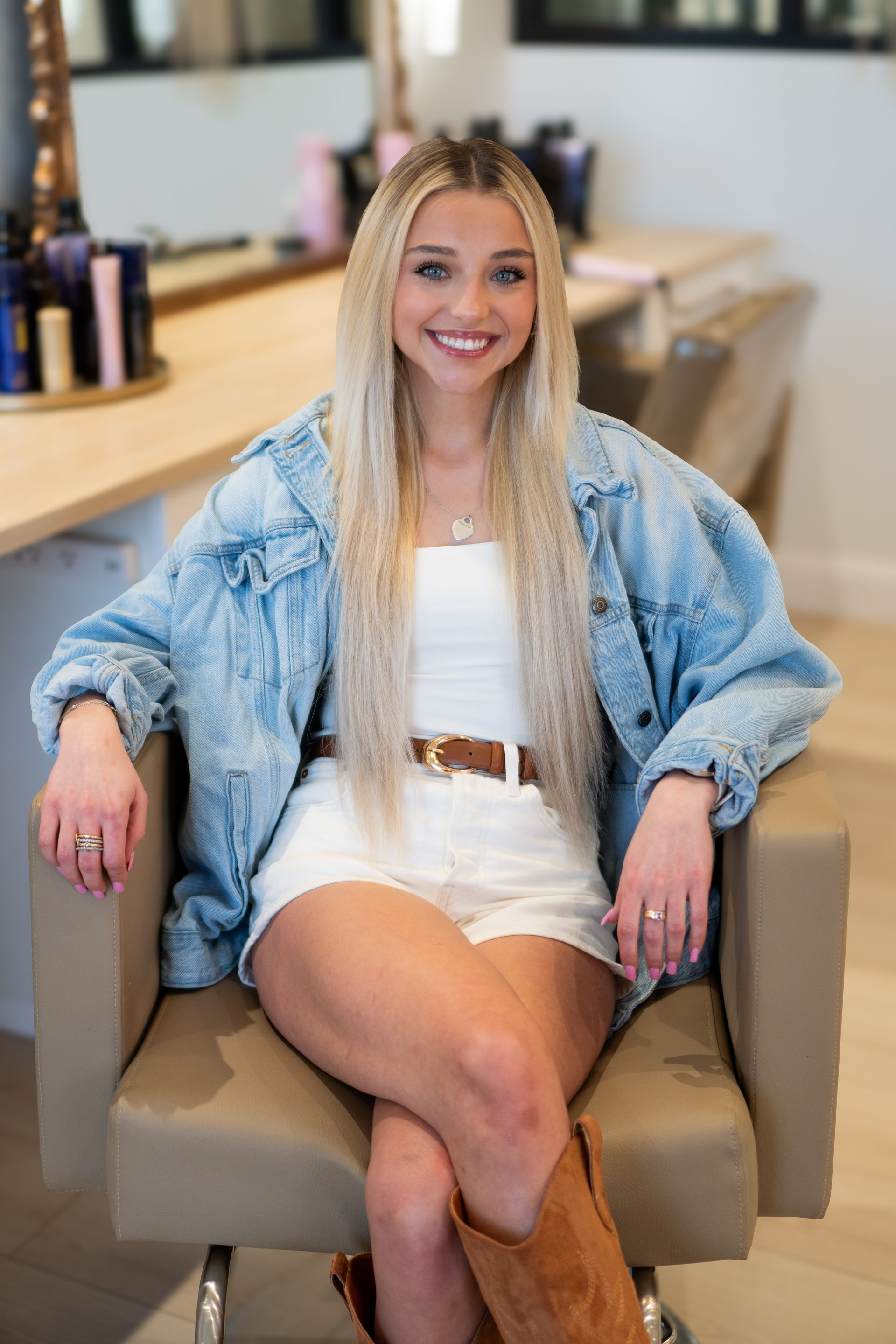 Blonde woman in denim jacket and white outfit sits in a chair in a salon.