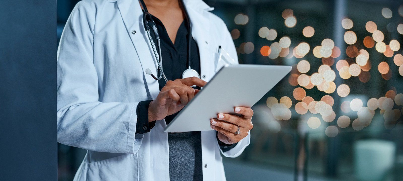 Medical professional in a lab coat, using a tablet computer. Blurry lights in the background.