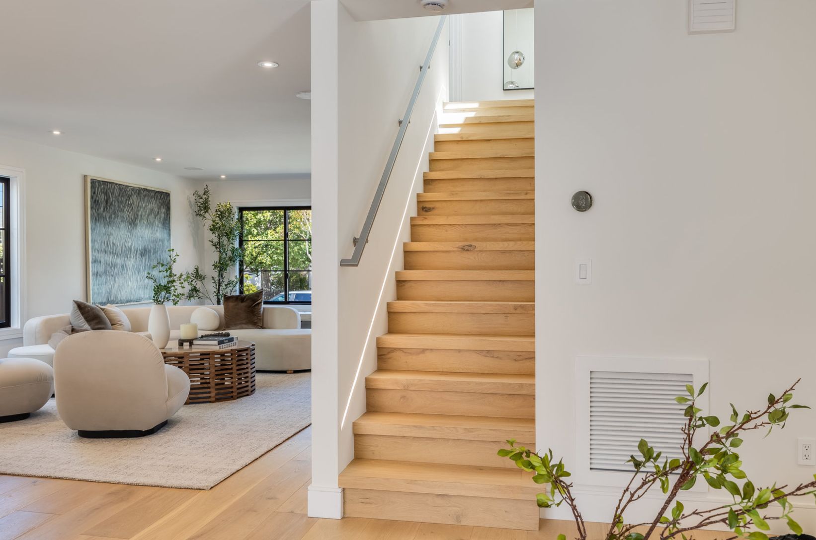 Interior view of a house with wooden stairs, light walls, and a living area with a sofa, rug, and artwork.