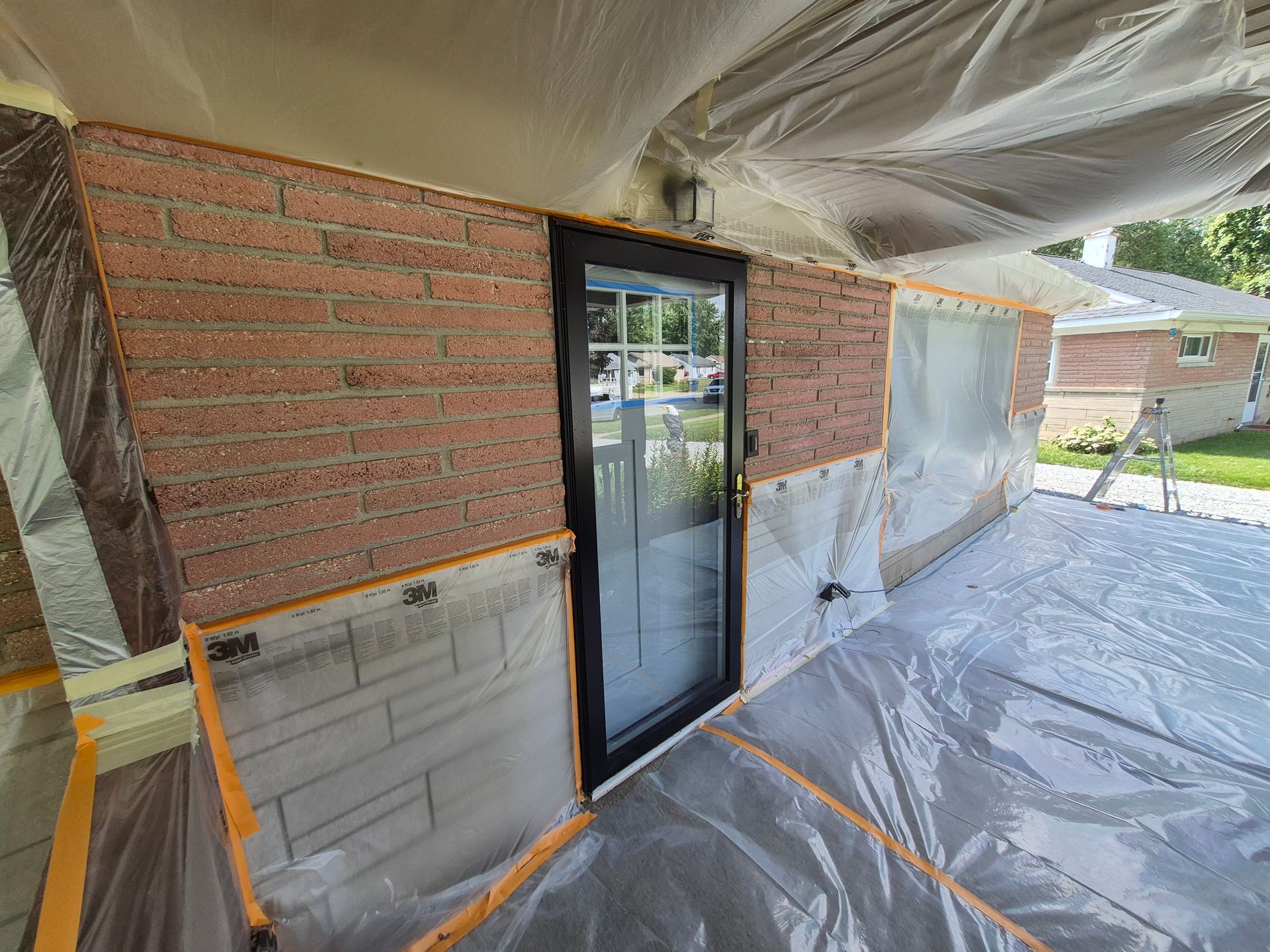 Exterior brick wall with a black-framed door, covered in plastic sheeting for painting.