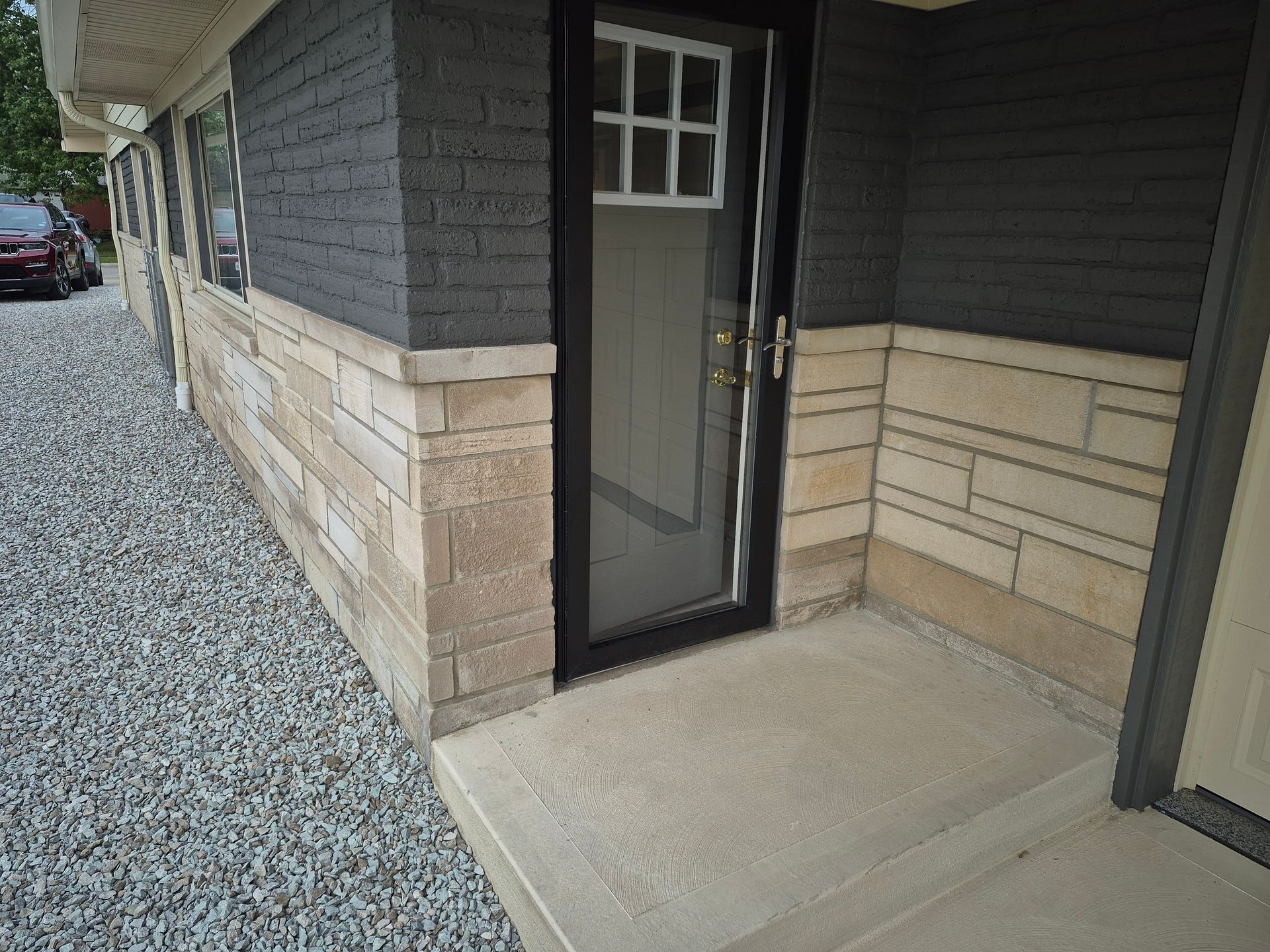 Entryway with stone veneer, dark siding, and black door. Concrete porch and gravel next to building.