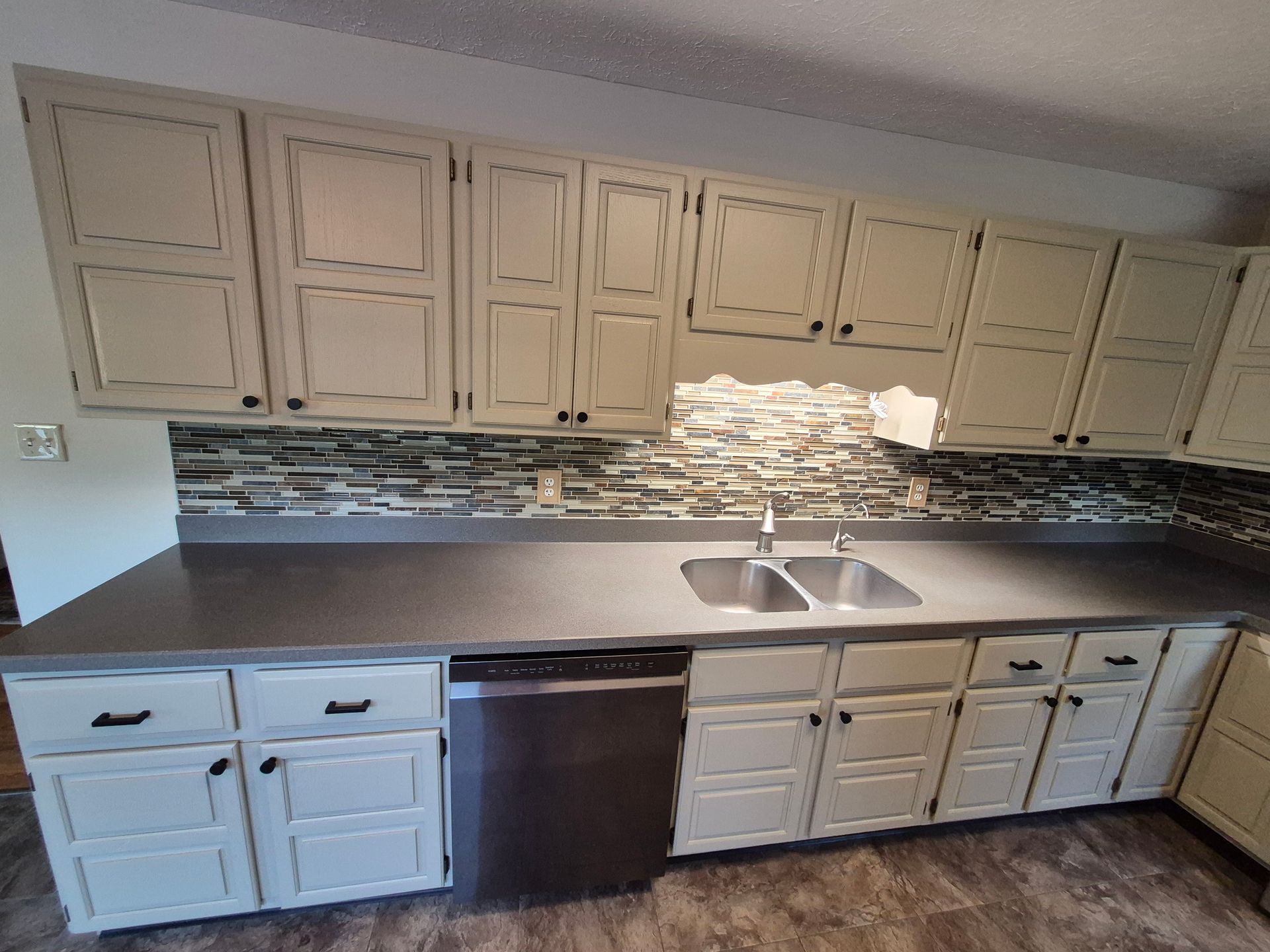 Cream-colored kitchen cabinets with dark hardware, gray countertop, stainless steel appliances, and mosaic tile backsplash.
