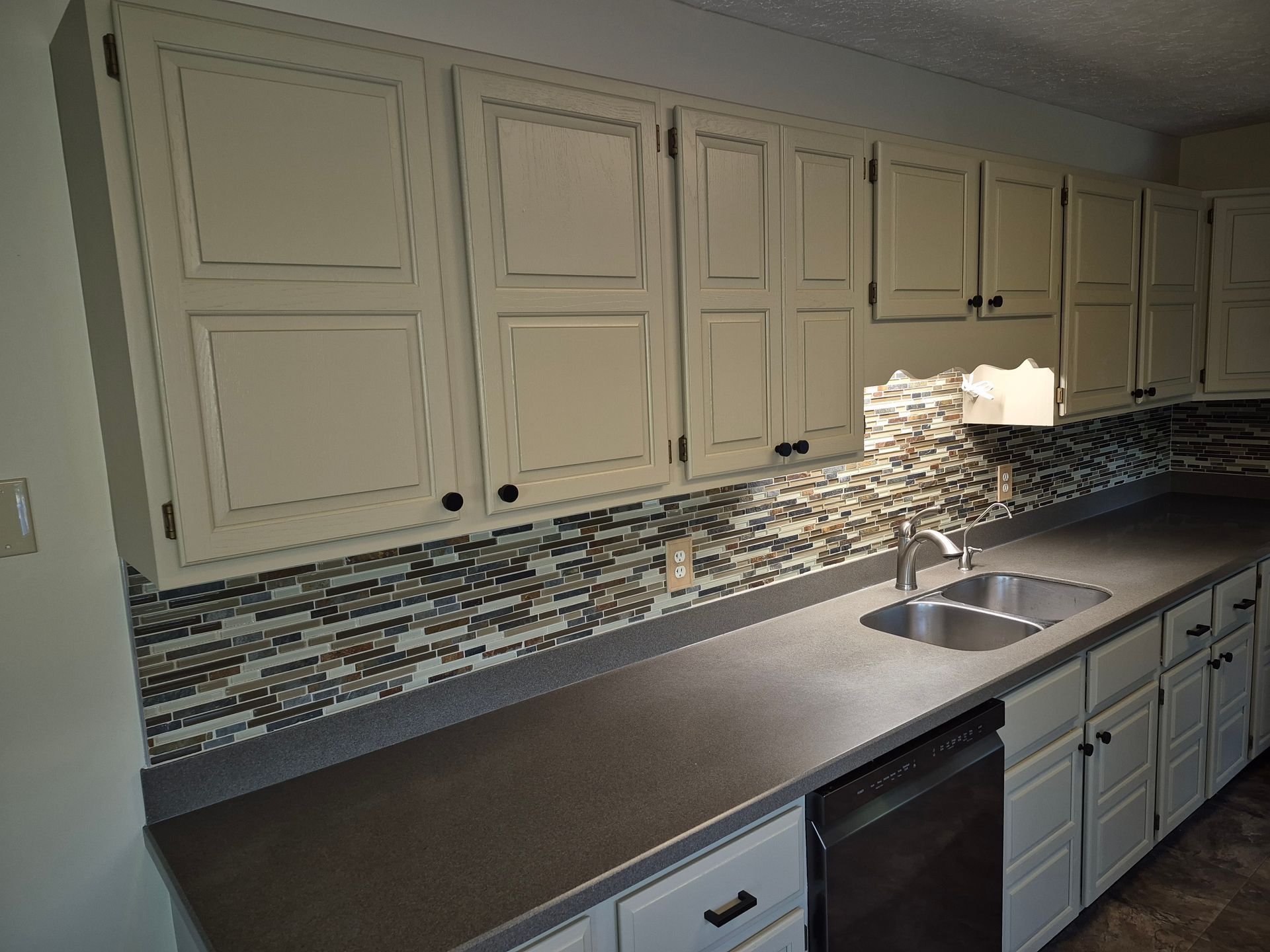 Kitchen with off-white cabinets, dark gray countertop, mosaic tile backsplash, and stainless steel sink.