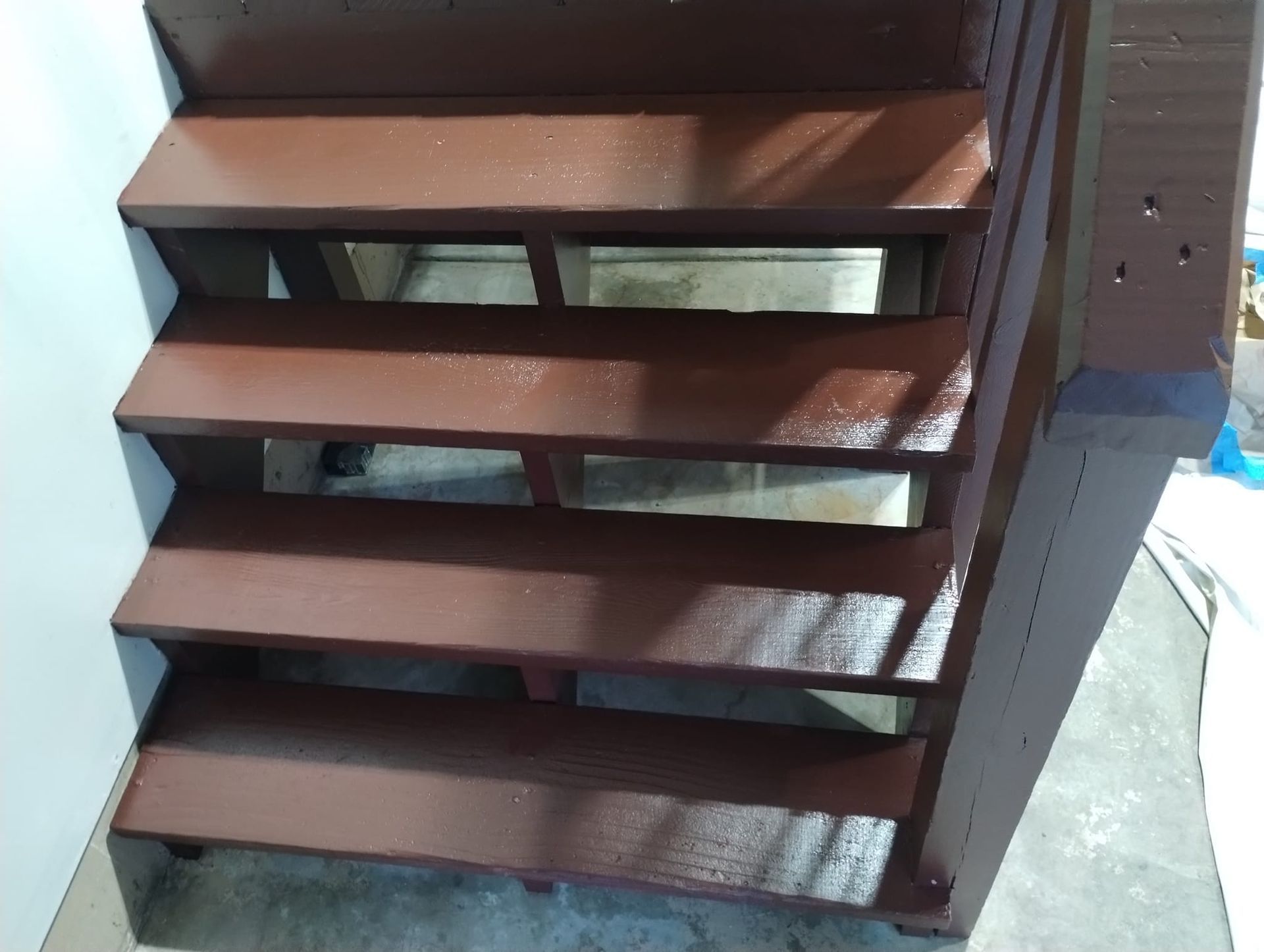 Brown wooden stairs with open risers, leading upwards against a white wall.