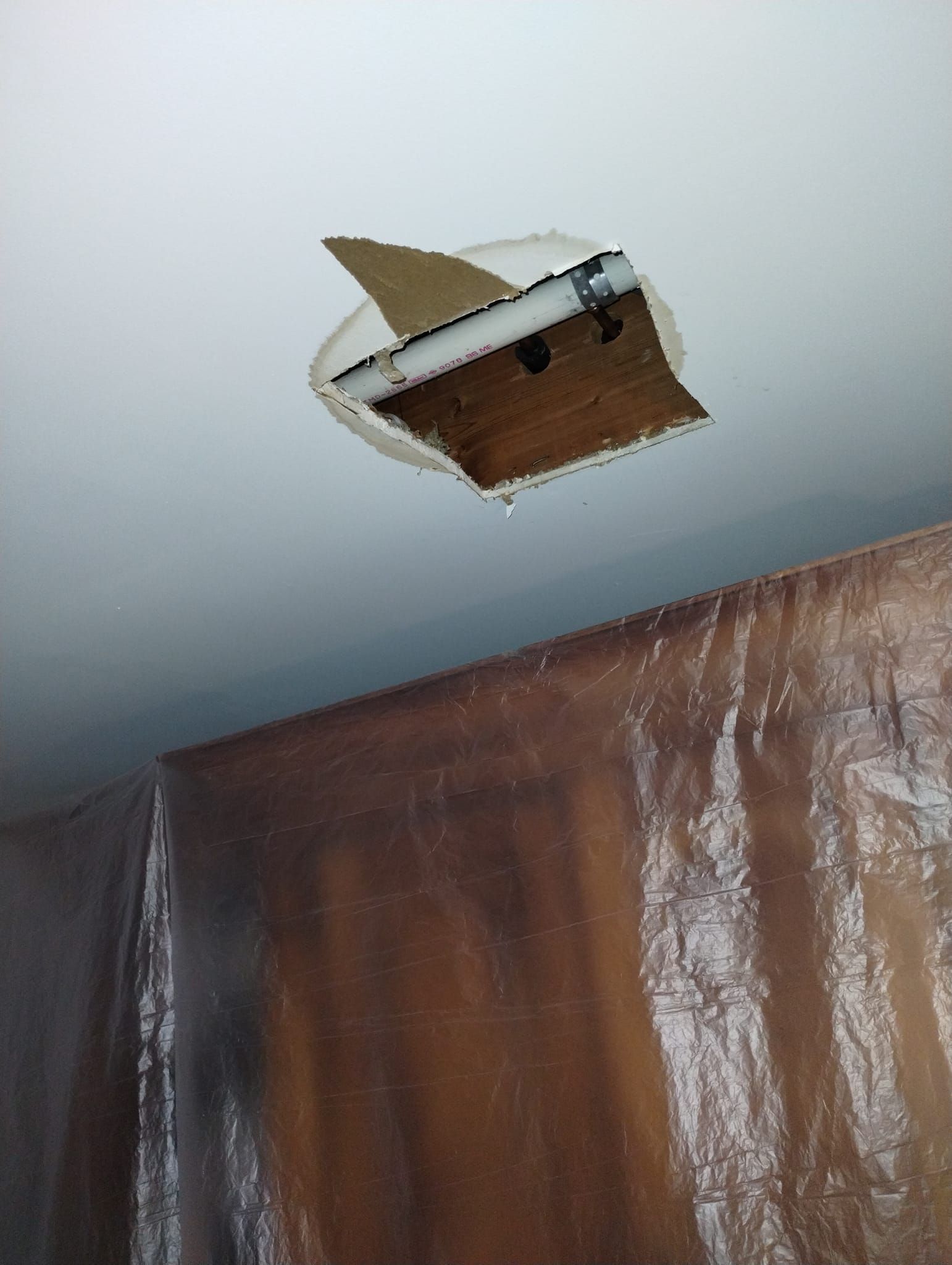 Hole in ceiling with exposed fixture, water dripping. Plastic sheeting covers a brown wall.