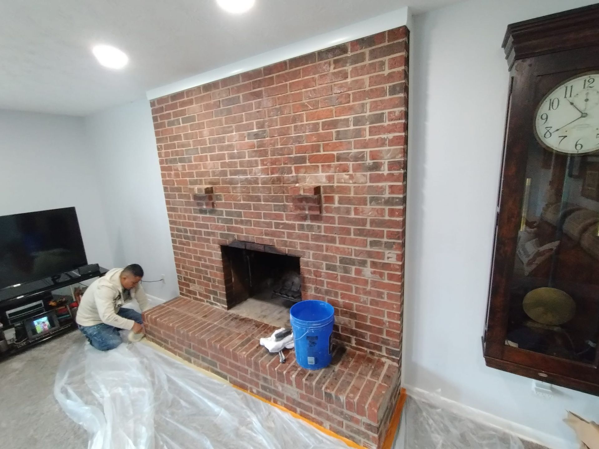 Man cleaning a red brick fireplace in a room with blue walls, a large clock, and a TV.