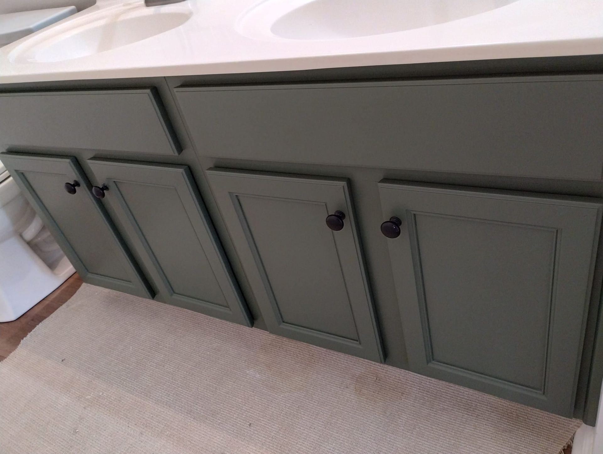 Green bathroom vanity with black knobs, white countertop, and a light-colored rug.