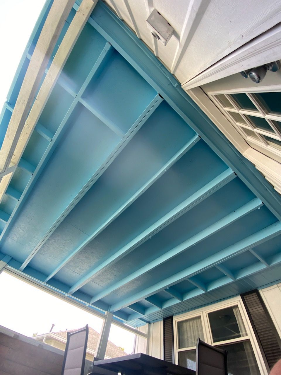 Blue-painted porch ceiling with supporting beams, attached to a white house with dark shutters.