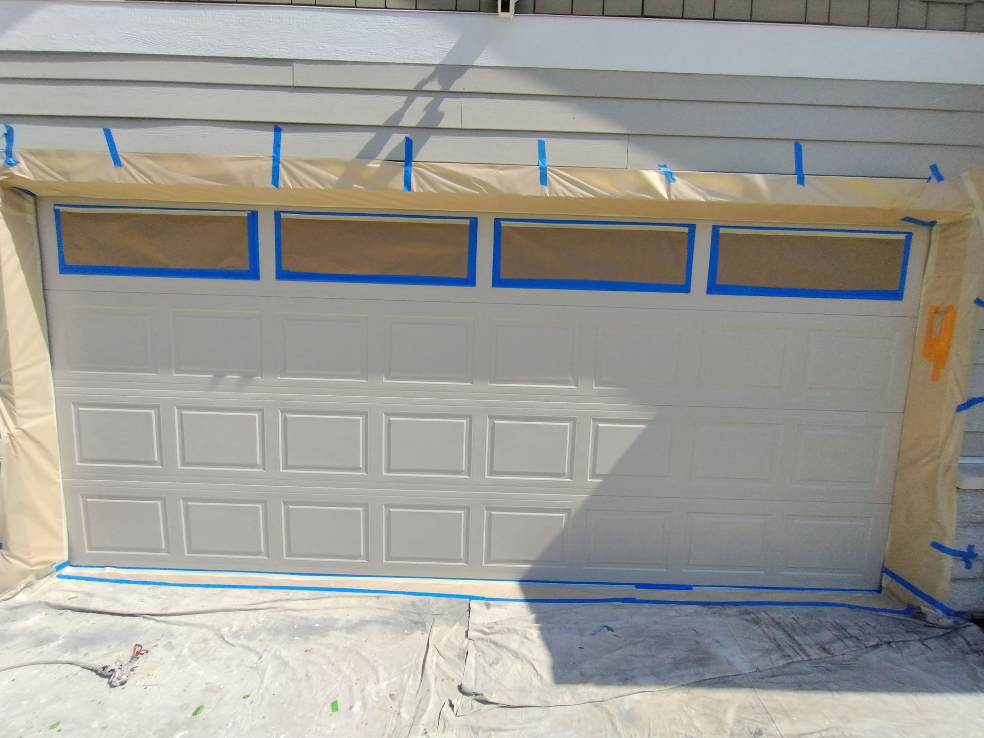 Garage door covered in tape, ready for painting. Beige door, light blue tape, brown paper, and gray siding.