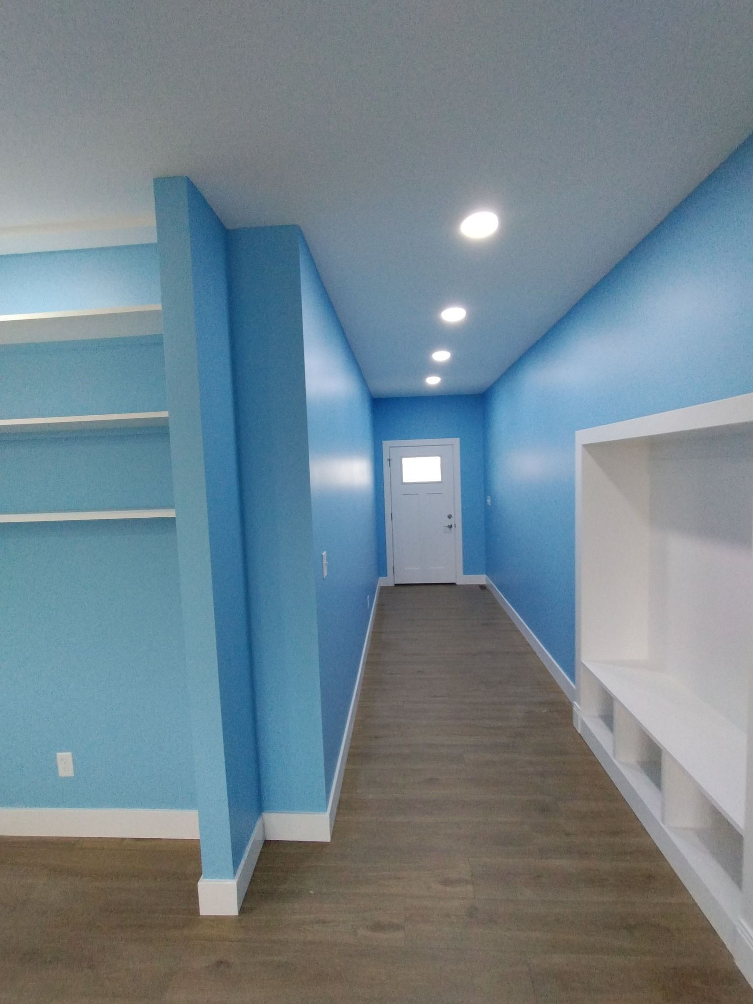 Long, narrow hallway painted light blue with white built-ins and recessed lighting. A white door is at the end.